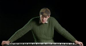 Sean Logan standing over a piano keyboard, arms wide, wearing a flat cap and looking at the camera