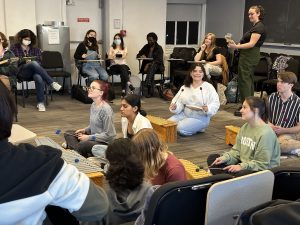 NYU Students sitting on the floor playing percussion instruments with students with guitars and iPads seated behind them