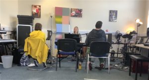 A group of three in a room. Two females, one in yellow, one in grey face their teacher in a stripey top sitting at a keyboard. They are all singing.