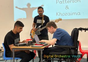 DMS Musician, Farhad wearing black is playing bass guitar. In front of him sitting at a desk, are two boys. One all in black on the left playing an iPad, and the other in blue on the right playing a laptop and keyboard.